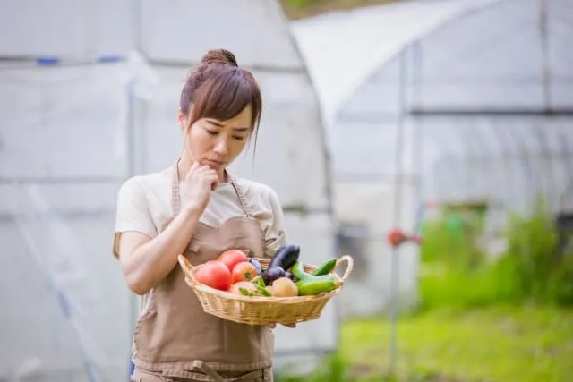 収穫した野菜をカゴに入れて持ちながら何かを考えている女性農家の写真