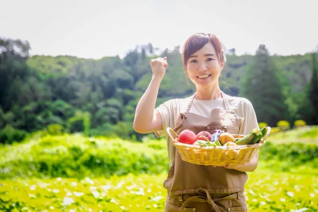 収穫した野菜を網カゴにいれて微笑みながら持つ女性農家の写真