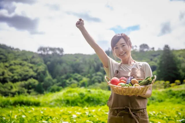 女性農家が明るい表情で収穫した野菜を片手に抱えながら拳を上げている写真